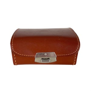 May include: A brown leather case with a silver metal clasp. The case has a stitched edge and is closed.