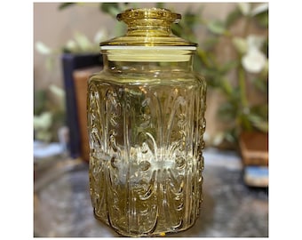 May include: A decorative amber glass jar with a lid. The jar has a square shape with ornate embossed floral patterns. The lid is also amber and has a decorative knob. The jar is likely used for storage or display.