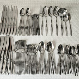 May include: A large set of stainless steel flatware featuring a polished finish and decorative embossed detailing on the handles. The collection includes various knives, dinner forks, salad forks, soup spoons, teaspoons, and a serving ladle.