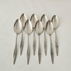 May include: A set of seven silver-toned metal spoons arranged in a cluster. Each spoon features a polished oval bowl and a slender handle decorated with an embossed floral motif near the base, showcasing a consistent design across all pieces.