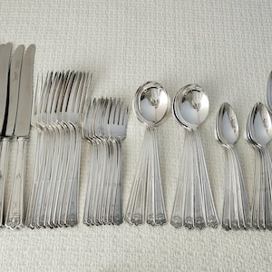 May include: A large set of silver-toned flatware including dinner knives, dinner forks, salad forks, soup spoons, teaspoons, and butter knives. Each piece features a slender handle with a decorative embossed pattern at the base.