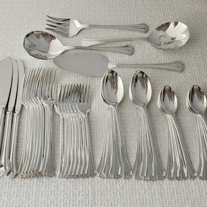 May include: A large set of polished silver-tone stainless steel flatware including dinner knives, forks, spoons, a cake server, and serving utensils. Each piece features a classic handle design with subtle vertical fluting and decorative detailing.