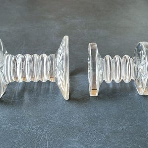May include: Two clear, cut-glass bottle stoppers. Each stopper has a decorative, circular top and a ribbed, cylindrical body. The stoppers are separated, showcasing their individual designs and potential use for wine or spirits.