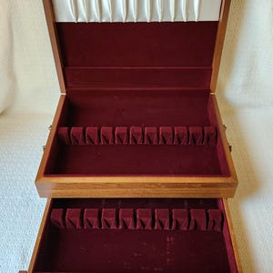 May include: A wooden flatware storage chest with a hinged lid and a pull-out drawer. The interior is lined with deep red velvet, featuring slotted dividers for silverware. The inside of the lid has a white pleated fabric panel.
