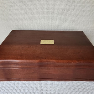 May include: A rectangular wooden box with a dark brown finish and a wavy, contoured front edge. Centered on the top lid is a small, rectangular brass-colored metal plate featuring an engraved monogram design with the letters G, B, and J.