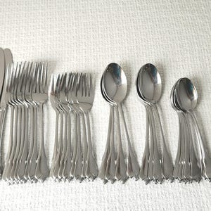 May include: A large set of polished stainless steel flatware including dinner knives, dinner forks, salad forks, soup spoons, and teaspoons. Each piece features a classic handle design with a subtle decorative ridge along the center.