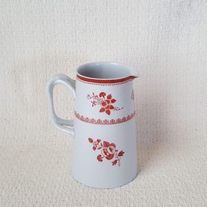 May include: A light gray ceramic pitcher with a handle and a spout. The pitcher is decorated with red floral patterns and a red band around the top. The pitcher is a classic design.