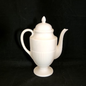 May include: A white ceramic teapot with a lid and a curved handle. The teapot has a fluted body and a pedestal base. The lid is topped with a decorative finial. The spout is curved and elegant. The teapot is set against a black background.