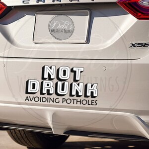 May include: White car bumper sticker with the text "NOT DRUNK AVOIDING POTHOLES" in a bold, 3D-style font. The sticker is on the rear of a white car, with the exhaust pipes visible.