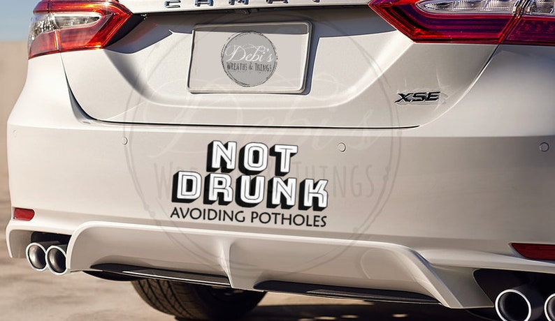 May include: White car bumper sticker with the text "NOT DRUNK AVOIDING POTHOLES" in a bold, 3D-style font. The sticker is on the rear of a white car, with the exhaust pipes visible.
