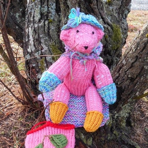 May include: A pink knitted teddy bear wearing a blue and purple hat with a flower on top. The bear is sitting on a brown tree trunk with a pink knitted bag with green leaves on the ground.