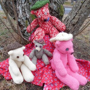 May include: Four knitted teddy bears with different coloured fur and hats. The bears are sitting on a red and white patterned blanket.