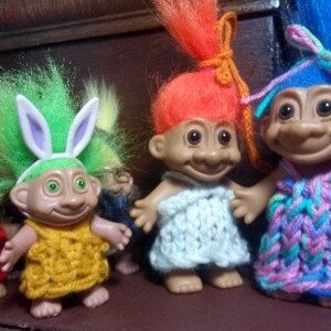 May include: Four troll dolls with colorful hair and knitted clothing. The dolls are wearing different outfits, including a green dress with bunny ears, a white dress, a pink and blue dress, and a red dress.