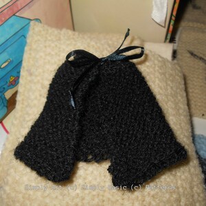 May include: A black knitted doll dress with a black velvet ribbon tied at the neck. The dress is on a cream-colored surface.