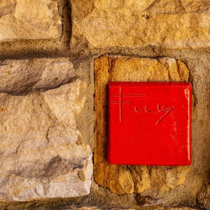 May include: A red square tile with the word "F.C.U." written in white on it, mounted on a stone wall.
