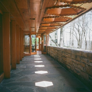 以下が含まれることがあります： 木製の壁と石の床を持つ、長く狭い廊下。窓から太陽光が差し込み、床に光と影のパターンを作っています。