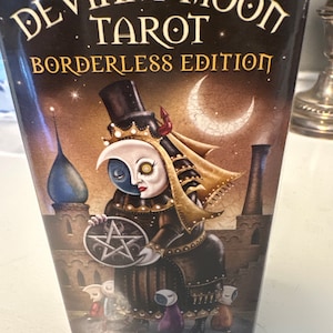 May include: A box for the Deviant Moon Tarot deck, featuring a borderless edition with a dark, whimsical illustration of a figure with a crescent moon head, holding a pentagram. The text "Deviant Moon Tarot Borderless Edition" is printed on the box.