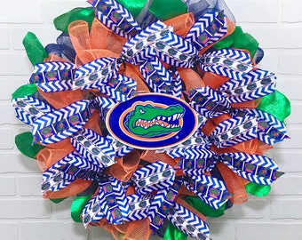 May include: A handmade orange, green, and blue wreath with a white and blue chevron ribbon and a blue and green alligator logo. The wreath is decorated with mesh and ribbon.