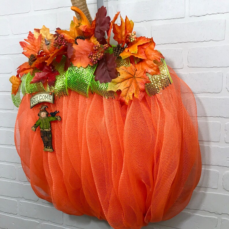 May include: Orange mesh pumpkin wreath decorated with autumn leaves, a green and gold ribbon, and a welcome sign featuring a scarecrow figure.