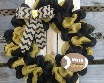 May include: A black and gold burlap wreath with a black and white chevron bow and a football-shaped ornament that says "Pittsburgh Steelers".