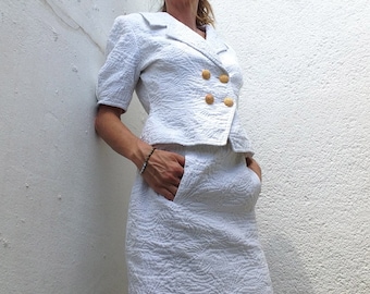 Puede incluir: Un traje blanco de dos piezas con textura, con una chaqueta corta y una falda. La chaqueta tiene cuello, cierre de doble botonadura y botones dorados. La falda tiene una silueta recta y bolsillos.