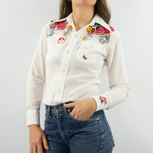 Può includere: Una camicia bianca a maniche lunghe con bottoni con ricami floreali colorati sul colletto e sulle spalle. La camicia ha una vestibilità rilassata e presenta un unico design a farfalla sulla tasca sul petto sinistro.