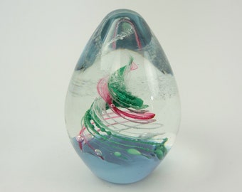 May include: A clear glass paperweight with a swirl of pink, green, and white glass inside. The paperweight has a light blue base.