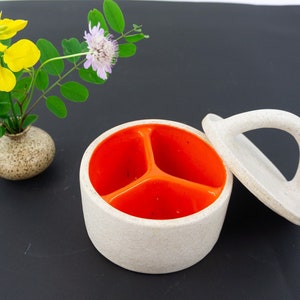 Puede incluir: Un recipiente de cerámica blanca con tapa y asa. El recipiente tiene tres compartimentos en el interior, cada uno pintado de un color naranja brillante. La tapa está abierta y el recipiente está sobre una superficie negra.