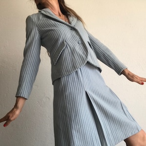 Puede incluir: Un traje azul claro de rayas finas con una chaqueta y una falda. La chaqueta tiene un cierre de un solo bot&oacute;n y la falda tiene un frente plisado.