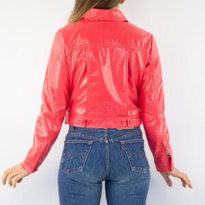 May include: A bright pink cropped jacket made of shiny faux leather. The jacket has a button-up front and long sleeves.