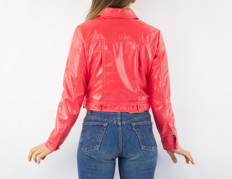May include: A bright pink cropped jacket made of shiny faux leather. The jacket has a button-up front and long sleeves.