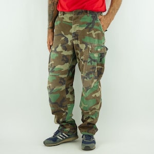 Könnte beinhalten: Tarnfarbene Cargohose in Grün-, Braun- und Beigetönen. Die Hose hat mehrere Taschen und eine lockere Passform. Das Model trägt ein rotes Shirt und blaue Turnschuhe. Die Hose ist ein klassischer Militärstil.