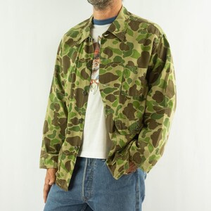 May include: A long-sleeve button-up shirt featuring a green, brown, and tan camouflage pattern. The shirt includes a pointed collar, a single chest pocket on the left side, and a lower pocket on the left side, both with button-down flaps.