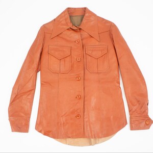 May include: An orange-brown leather shirt with a pointed collar, two chest pockets, and button closures. The long-sleeved shirt has buttoned cuffs and a curved hemline. The shirt is displayed on a white background.