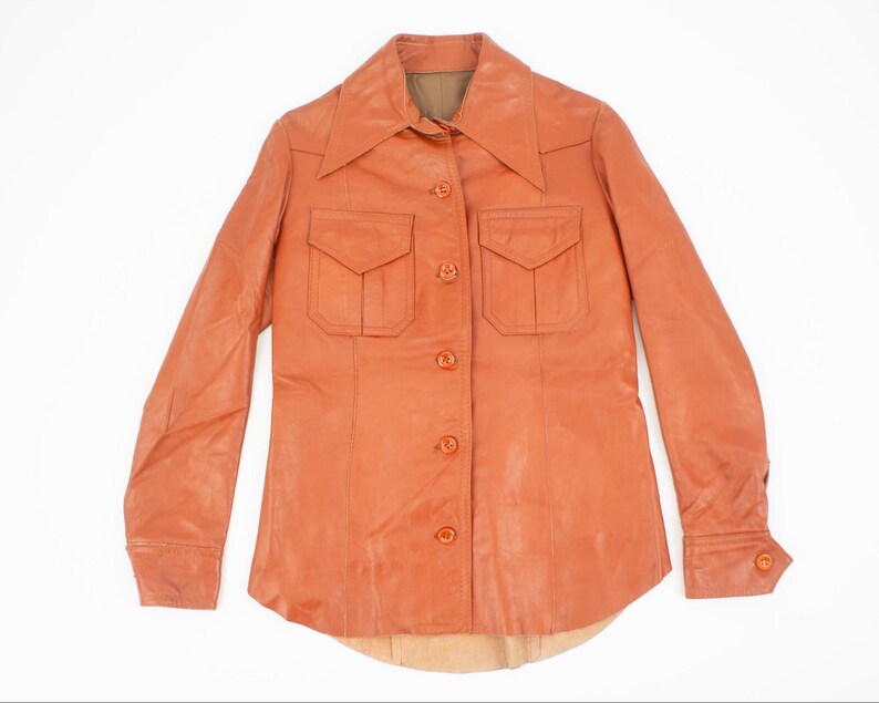 May include: An orange-brown leather shirt with a pointed collar, two chest pockets, and button closures. The long-sleeved shirt has buttoned cuffs and a curved hemline. The shirt is displayed on a white background.