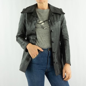 May include: Black leather jacket with a notched collar and button closure. The jacket is worn over a gray t-shirt and blue jeans. The jacket has a tailored fit and a classic design.