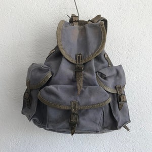 May include: A gray canvas backpack with brown leather straps and buckles. The backpack has two large pockets on the front and a smaller pocket on the top.