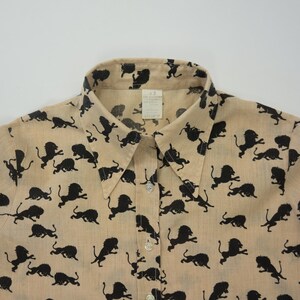 May include: A beige button-up shirt with a black lion print. The shirt has a collared neckline and short sleeves.