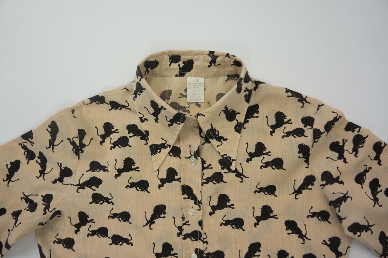 May include: A beige button-up shirt with a black lion print. The shirt has a collared neckline and short sleeves.