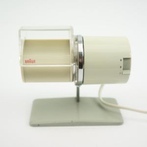 Könnte beinhalten: Vintage elektrische Kaffeemühle von Braun in Off-White und transparentem Kunststoff. Die Mühle hat einen zylindrischen Körper mit einem transparenten Oberteil, einem silbernen Metallring und einem grauen Sockel. Das Braun-Logo ist sichtbar.