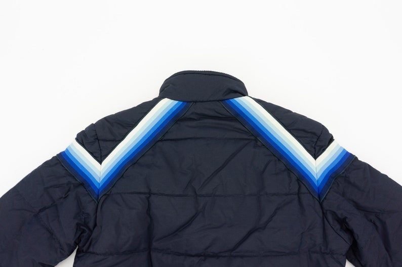 May include: A navy blue puffer jacket with a chevron pattern on the shoulders and sleeves. The chevron pattern is made up of white, light blue, and dark blue stripes.