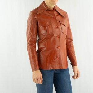 May include: A vintage, rust-colored leather shirt with a collared neckline, two chest pockets, and button closures. The long-sleeved shirt is paired with blue jeans. The shirt has a classic, retro style.