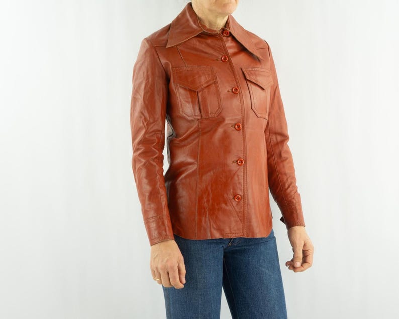 May include: A vintage, rust-colored leather shirt with a collared neckline, two chest pockets, and button closures. The long-sleeved shirt is paired with blue jeans. The shirt has a classic, retro style.