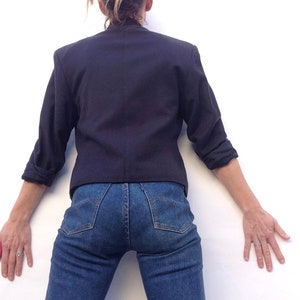 Pode incluir: Uma pessoa vestindo um blazer azul escuro e jeans azul. O blazer tem mangas compridas e gola.