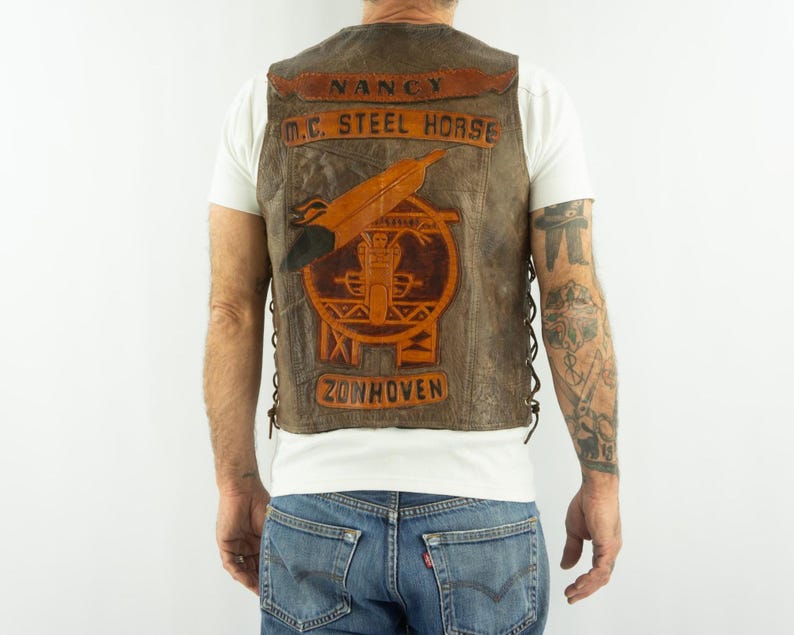 May include: Brown leather vest with the text "NANCY M.C. STEEL HORSE ZONHOVEN" and a motorcycle emblem. The vest has a distressed look with a lace-up side design.