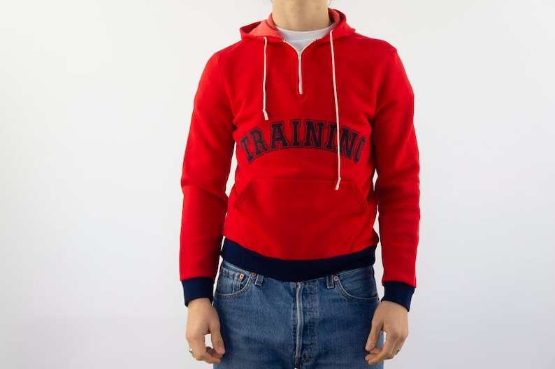 K&ouml;nnte beinhalten: Roter Kapuzenpullover mit dunkelblauem Saum und dem Wort "TRAINING" in einem dunkelblauen Bogen auf der Vorderseite.