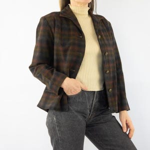 Op de afbeelding: Een bruine, groene en grijze geruite blazer met gouden knopen. De blazer is dichtgeknoopt en heeft een klassieke kraag. Het model draagt een beige coltrui en een grijze jeans.