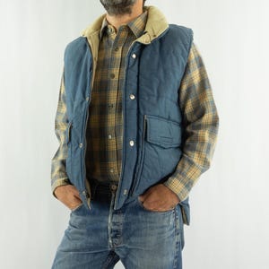Könnte beinhalten: Eine blaue Steppweste mit beigem Cordkragen wird über einem karierten Button-Down-Hemd und einer blauen Jeans getragen. Die Weste hat zwei Vordertaschen und ist zugeknöpft. Das Hemd ist gelb, braun und grau kariert.