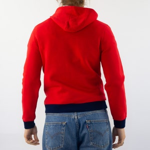 K&ouml;nnte beinhalten: Ein roter Kapuzenpullover mit einem marineblauen Saum um die Kapuze und den unteren Rand des Pullovers. Der Pullover wird von einer Person mit langen blonden Haaren getragen.