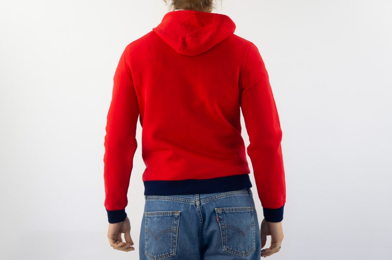 K&ouml;nnte beinhalten: Ein roter Kapuzenpullover mit einem marineblauen Saum um die Kapuze und den unteren Rand des Pullovers. Der Pullover wird von einer Person mit langen blonden Haaren getragen.